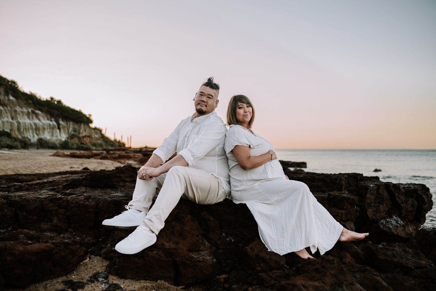 A couple in white outfits sits back-to-back on coastal rocks during sunset at the beach taken by Melbourne maternity photographer.