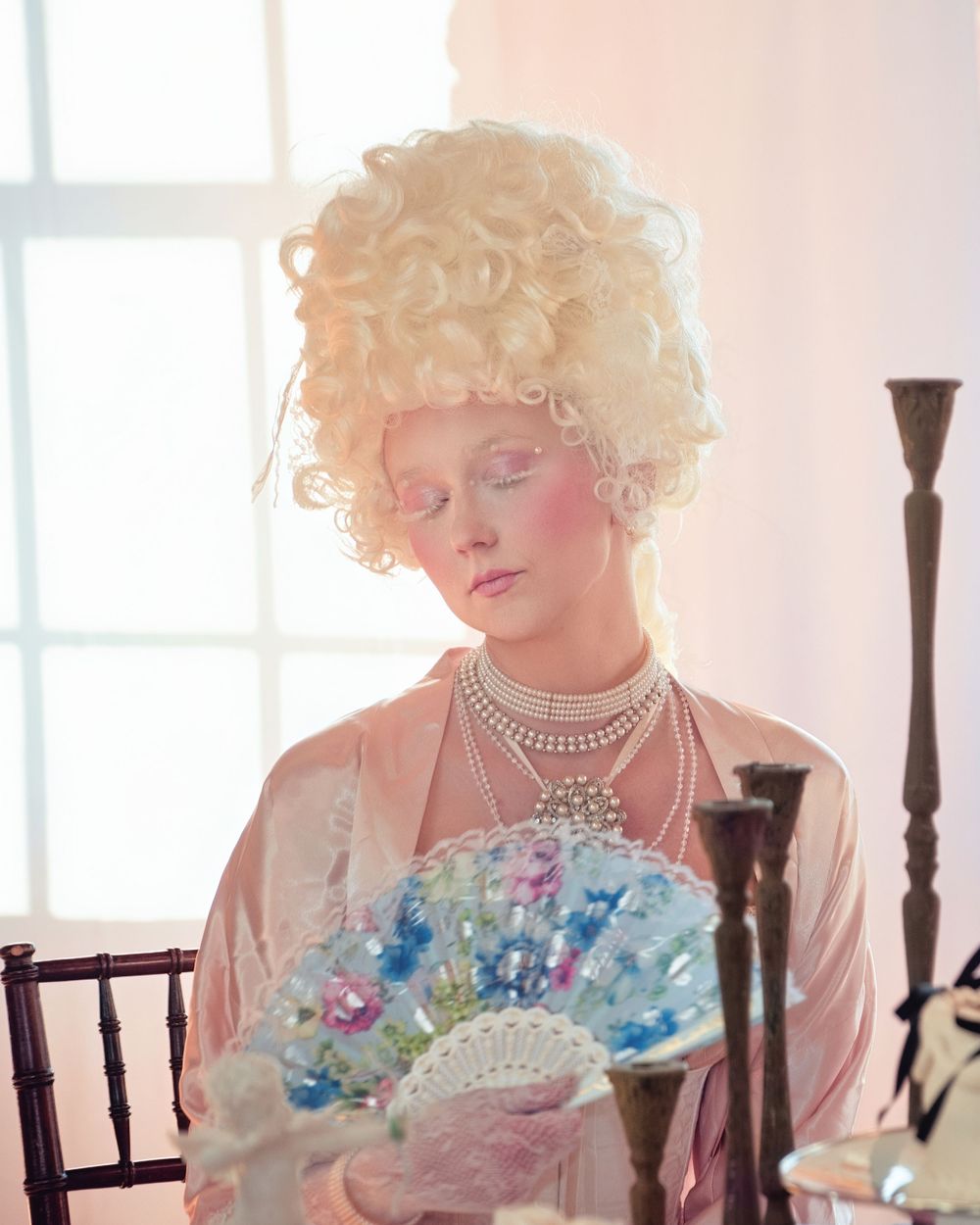 A person in a Marie Antoinette style white powdered wig and pearls holds a delicate fan against a soft pink background.