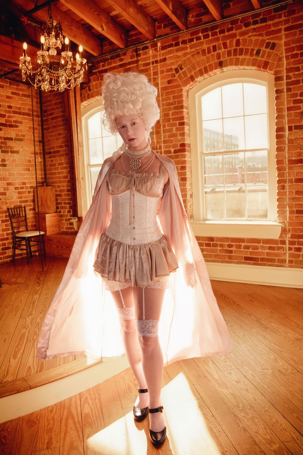 Victorian-inspired fashion with corset, flowing cape and stockings in a rustic loft space with exposed brick and chandeliers.