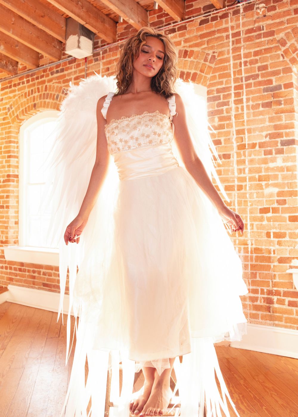 Fashion photo in white dress against exposed brick wall with natural light streaming in through windows on hardwood floor.