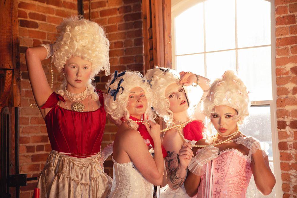A group in baroque-style costumes with powdered wigs pose in a room with exposed brick walls and arched windows.