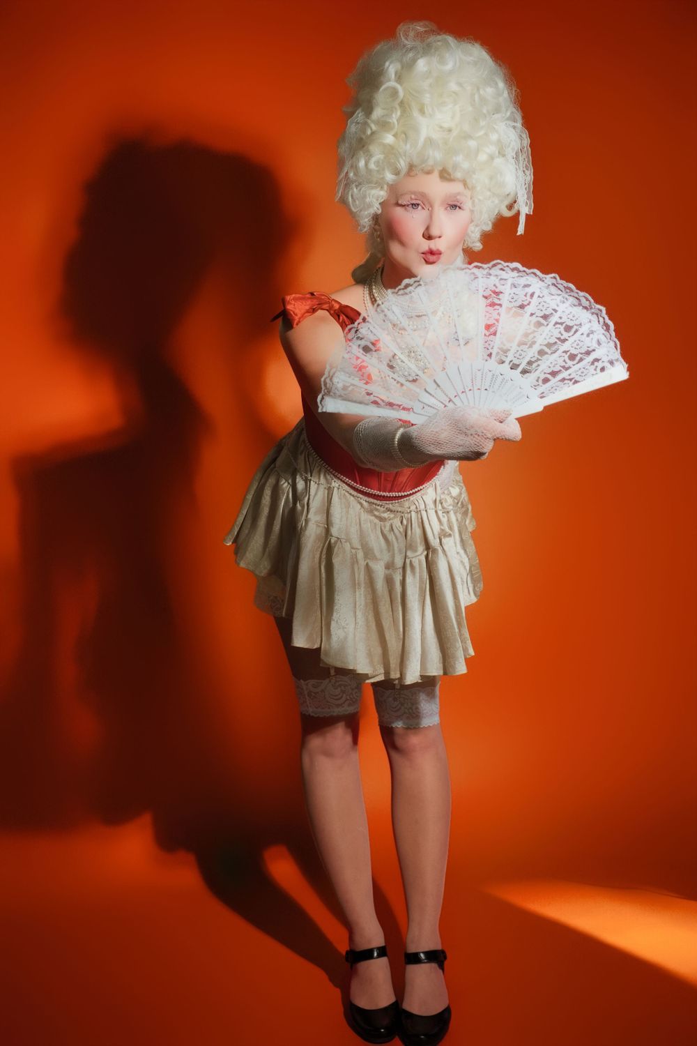 Figure in a white powdered wig and period dress holds a lace fan against an orange backdrop with dramatic shadow.