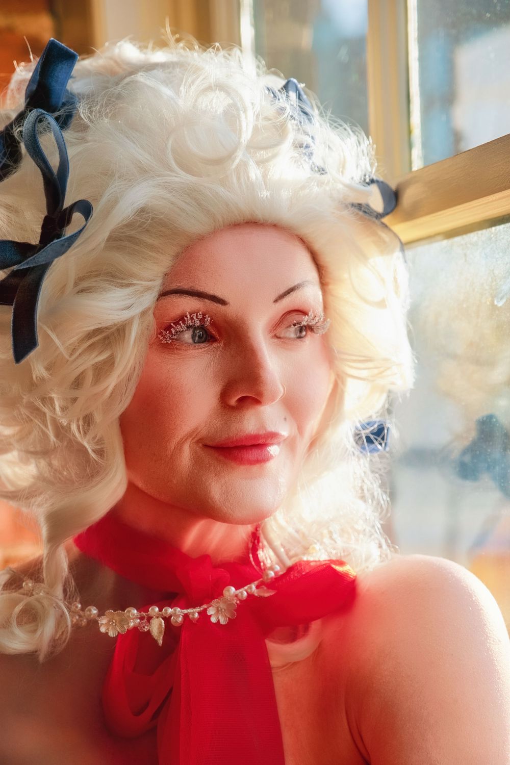Close-up portrait showing dramatic platinum blonde curls, bright red neckline, and delicate white lace details in soft lighting.