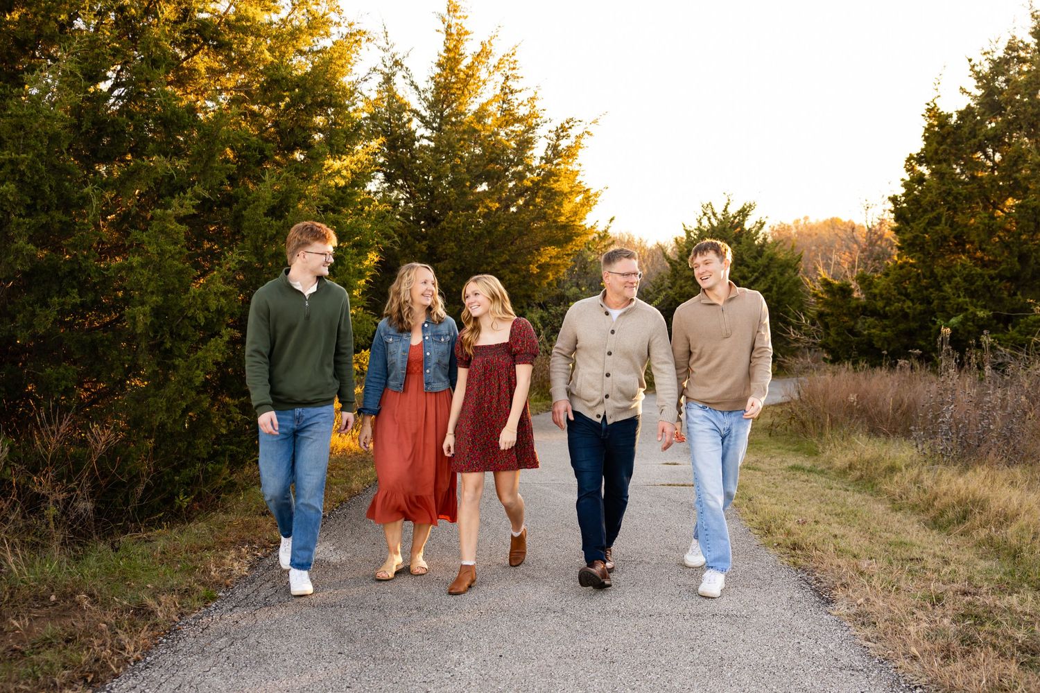 A group of people taking a sunset walk along a gravel path with evergreen trees and golden light in the background.