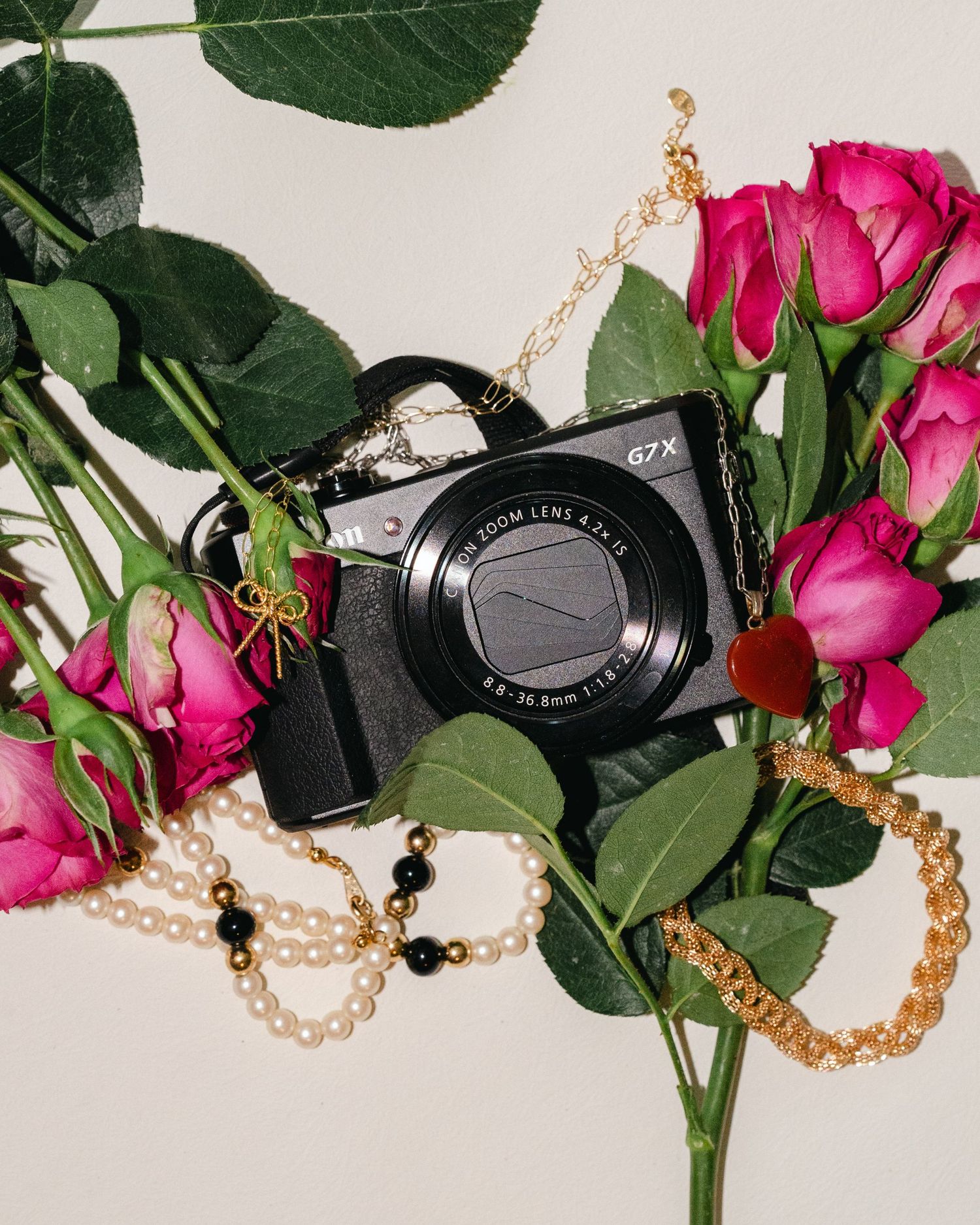 Black digital camera surrounded by vibrant pink roses and pearl necklaces on white backdrop.