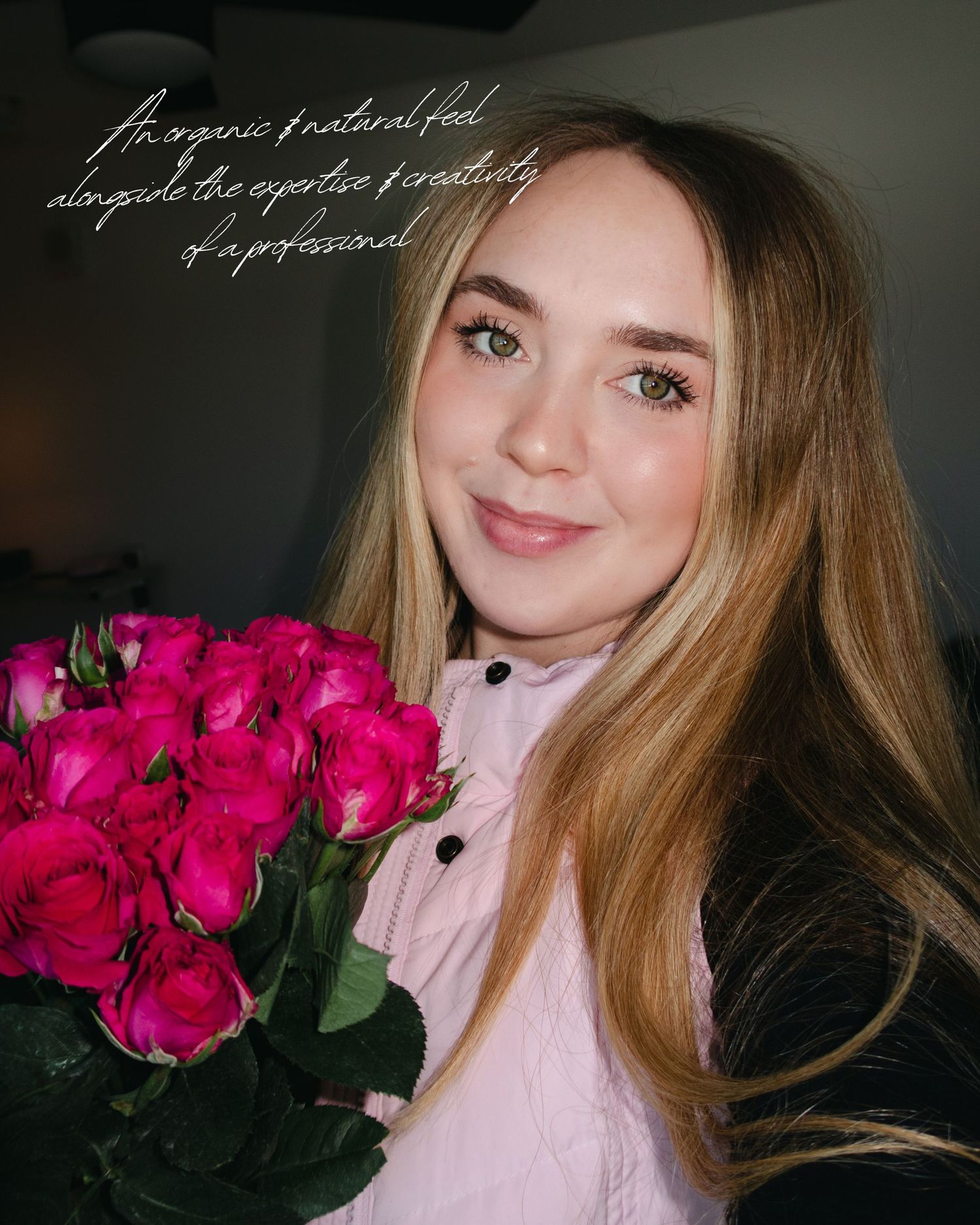 Person wearing pink button-up blouse holds bright magenta roses while smiling against dark background.