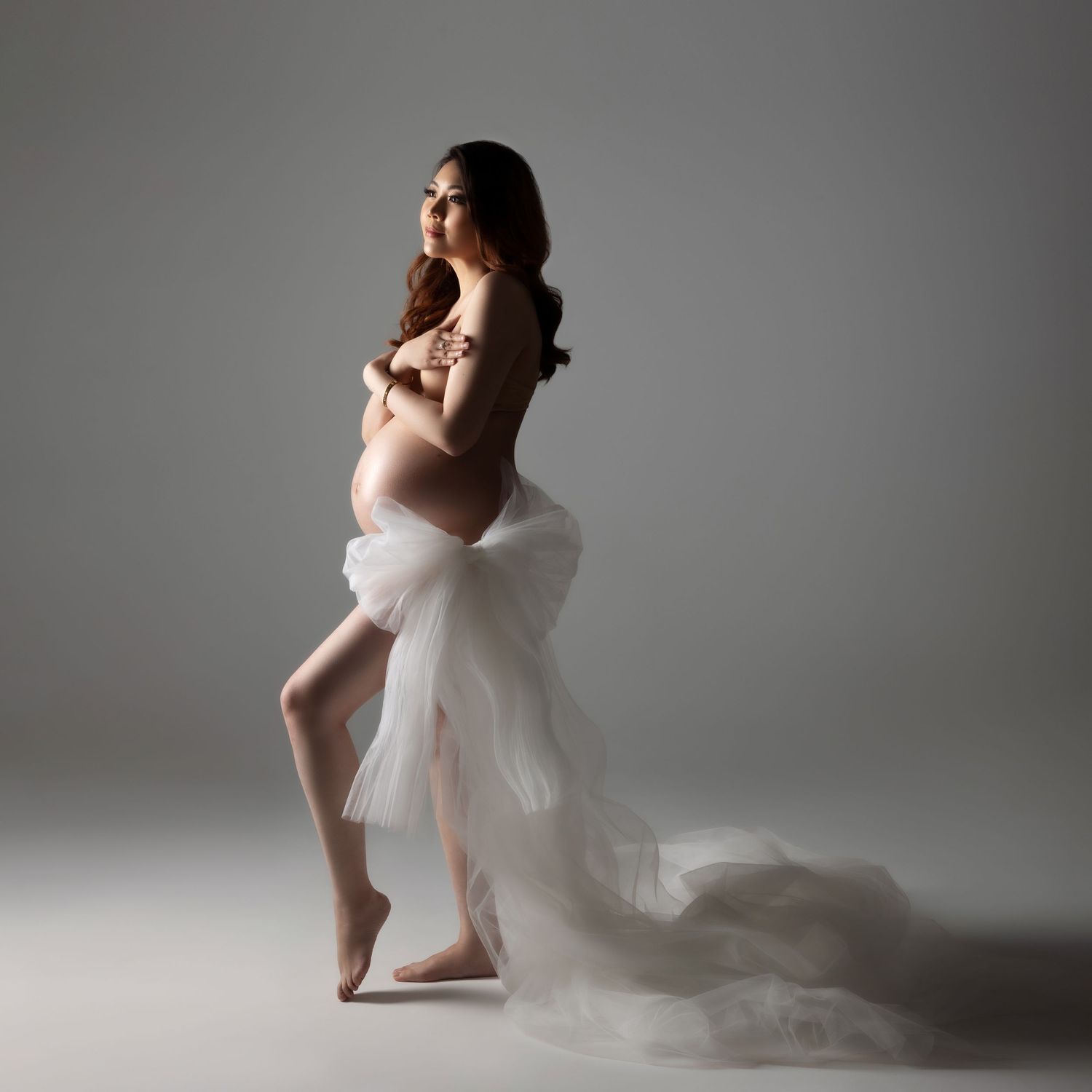Artistic maternity photo with flowing white fabric draping around an expectant mother in a minimalist studio setting.