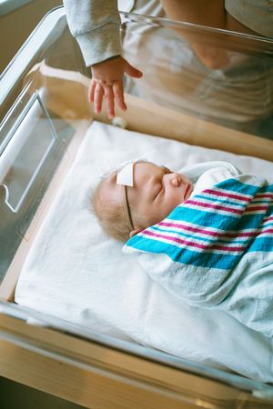 A newborn baby rests peacefully in a hospital bassinet wrapped in a striped blanket.