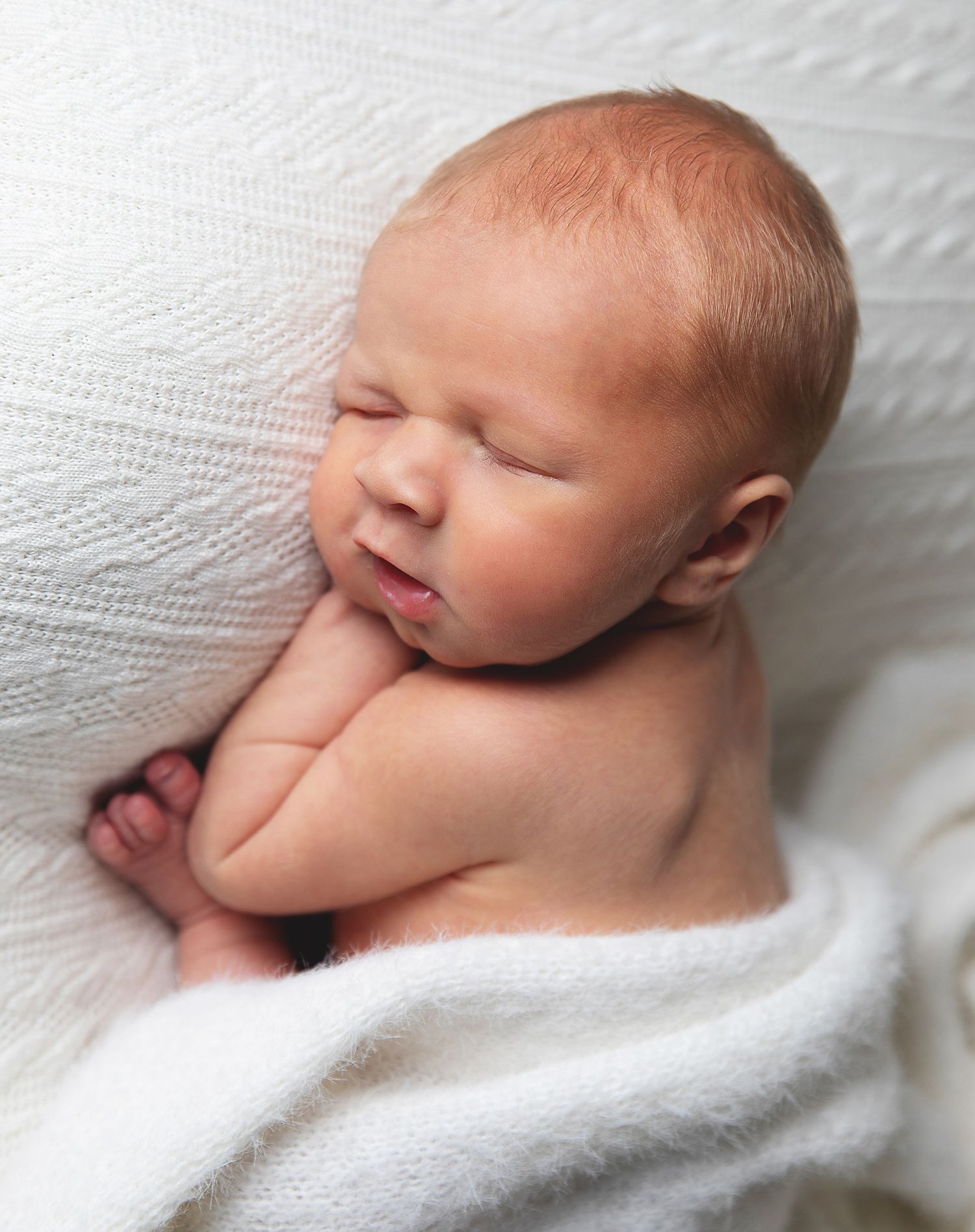 Peaceful sleeping newborn baby wrapped partially in soft white blanket laying on textured white surface. During a newborn session in Bexley