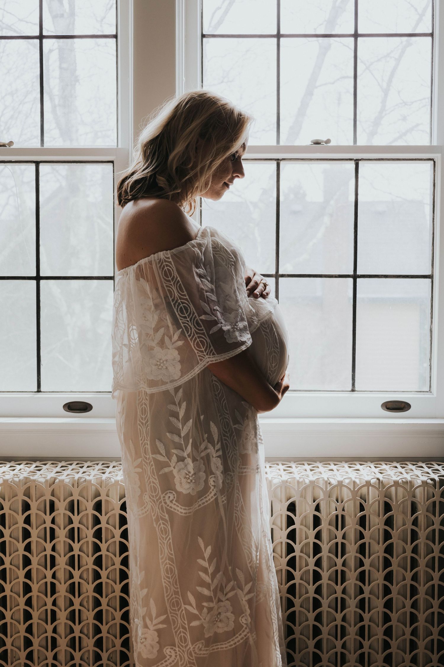 A silhouetted figure in a delicate lace gown stands near a window with intricate floral details during pregnancy.