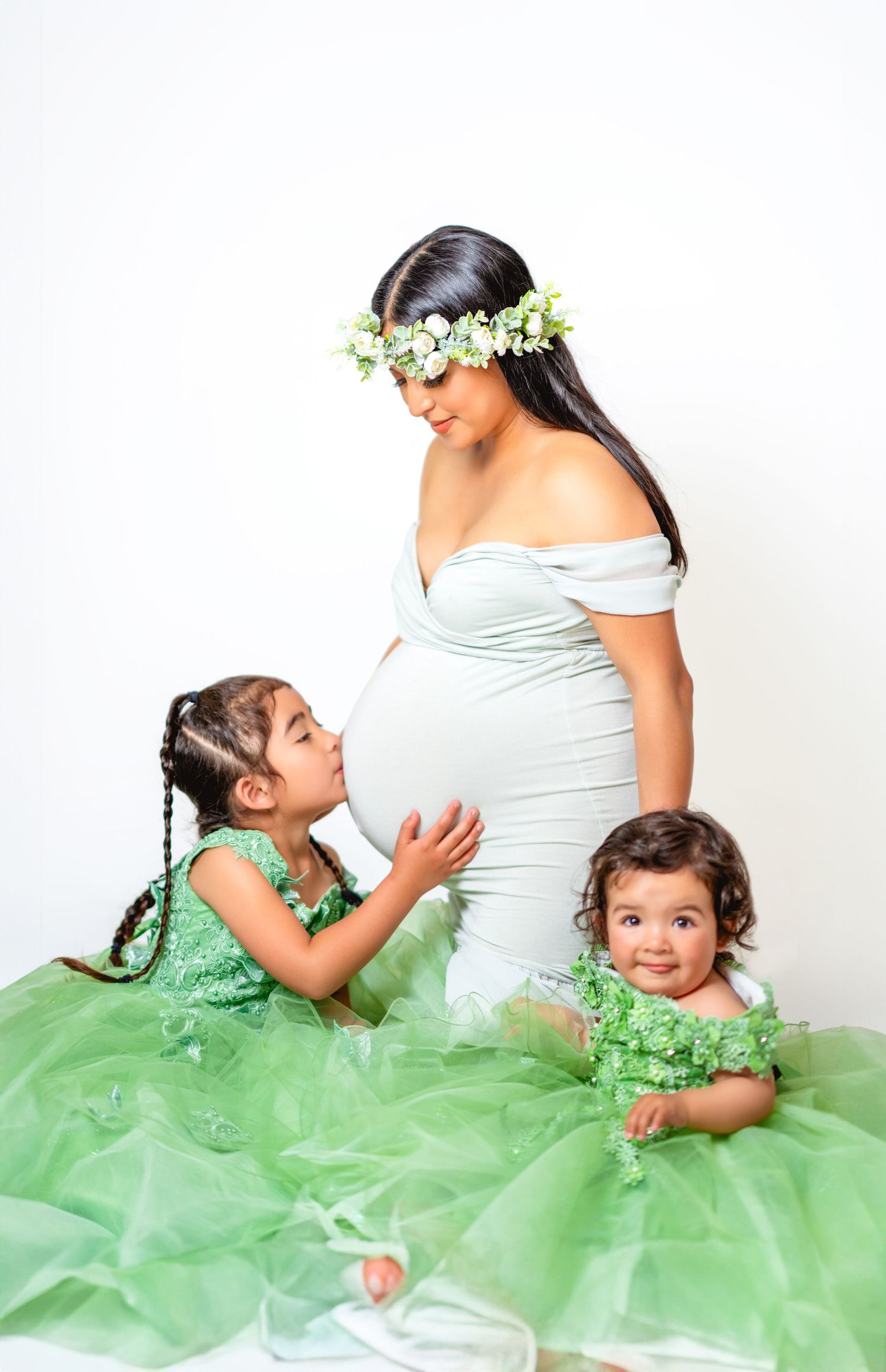 A pregnant woman in white dress and flower crown poses with two young children wearing matching green tulle dresses.