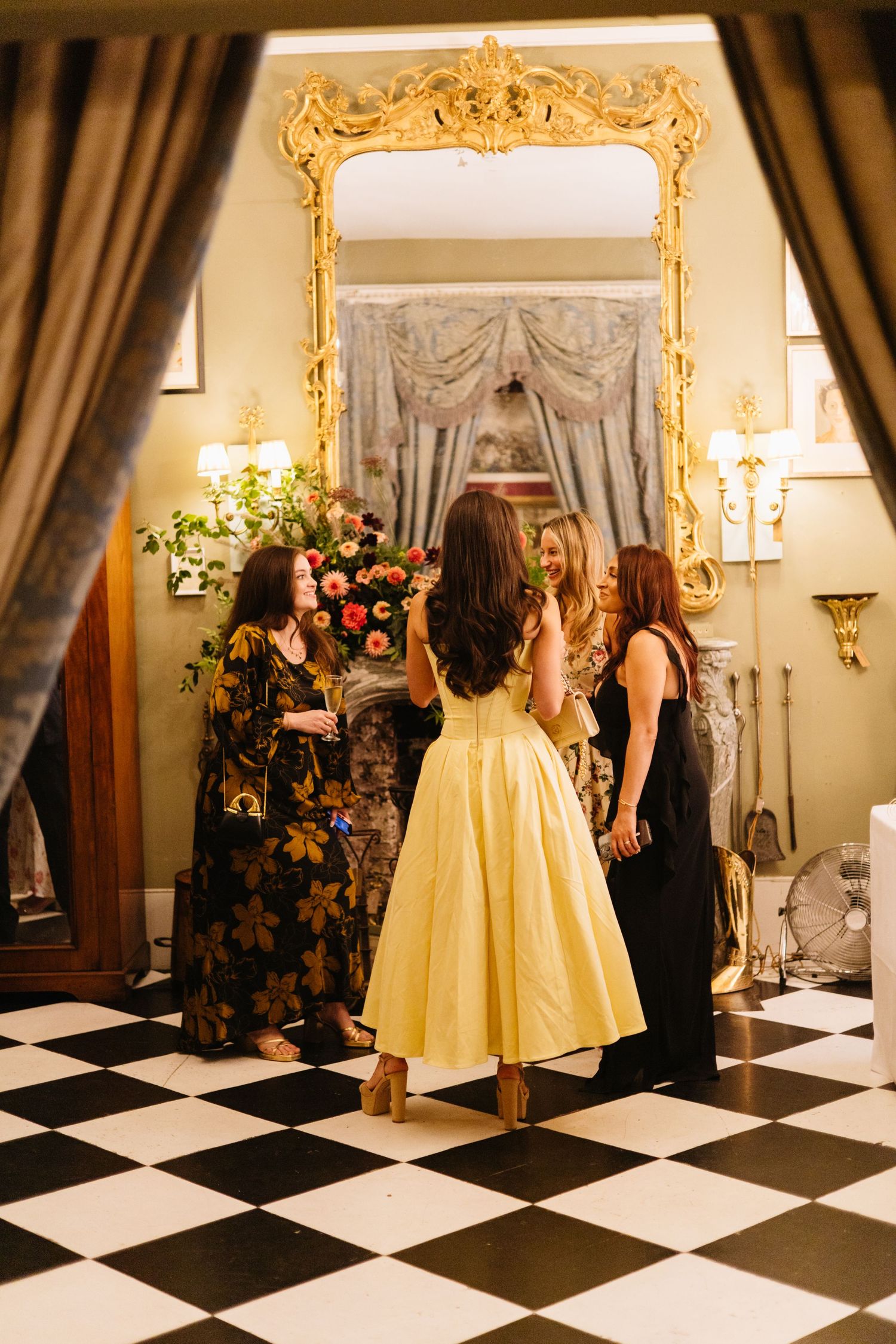 Elegantly dressed guests gather by an ornate gold mirror in a room with checkered floors.