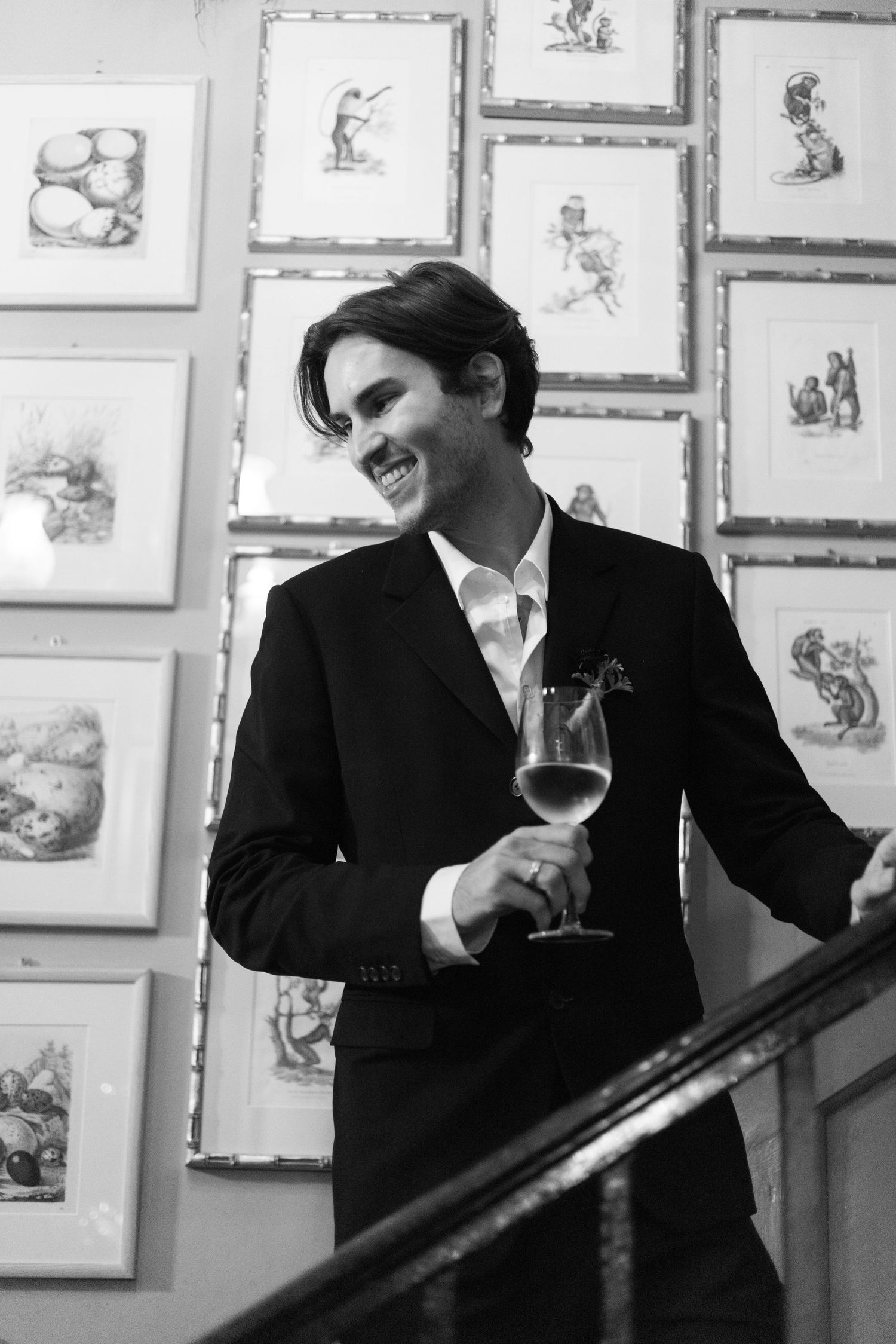 Person in formal attire holding wine glass against wall of framed artwork.