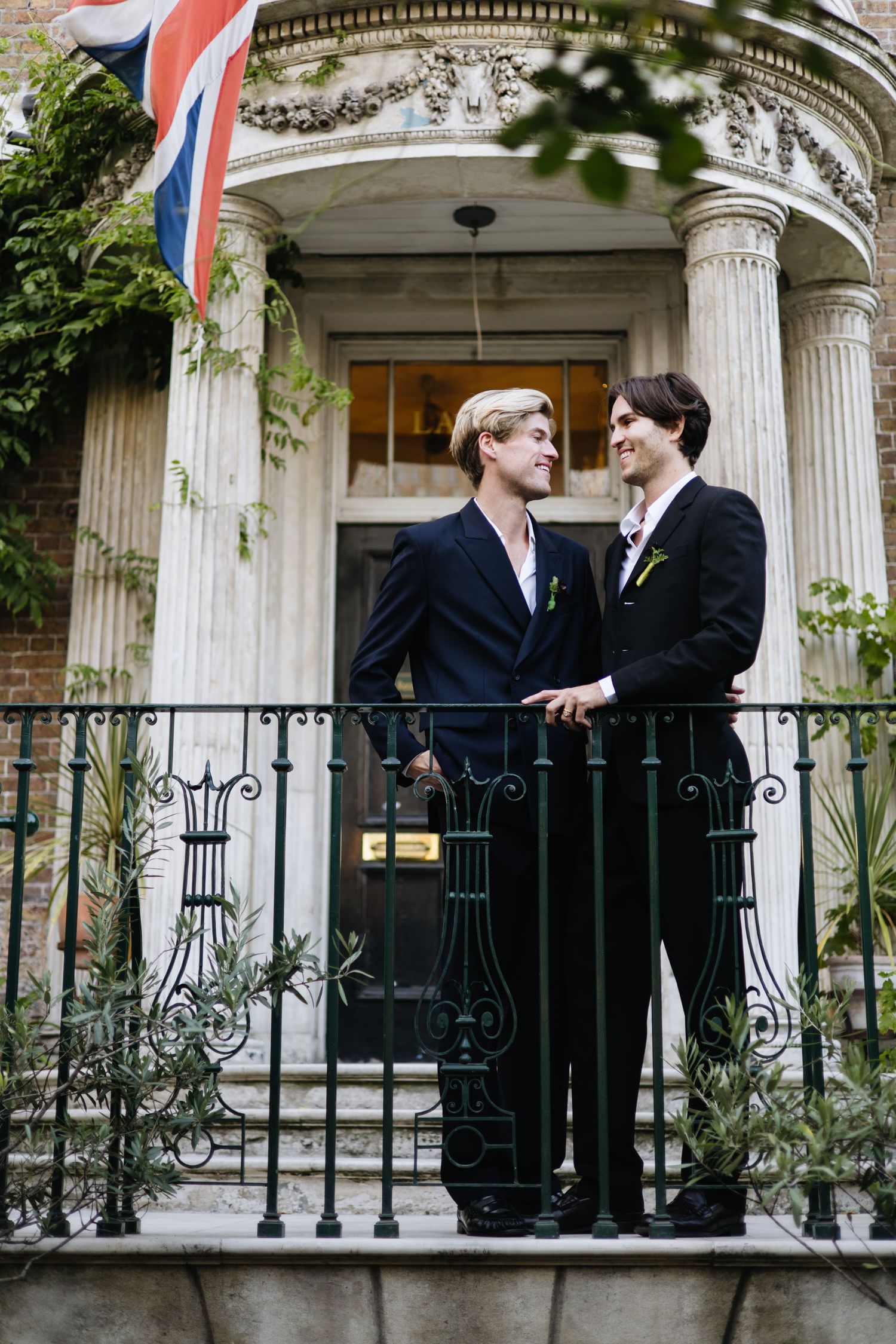 Two people in formal attire standing on a columned balcony with wrought iron railings.