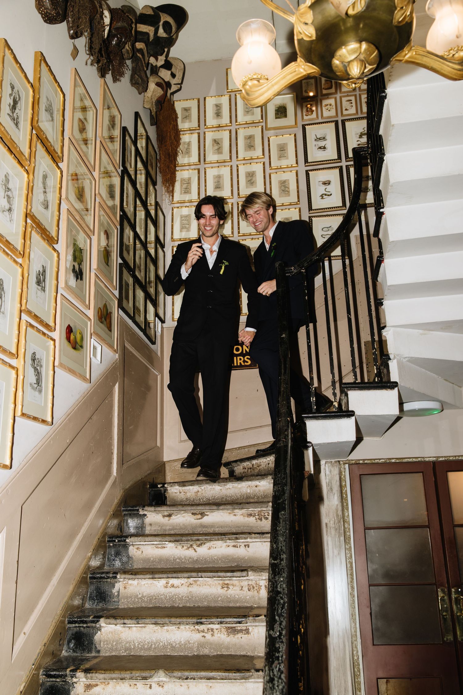 Two people in formal black attire descending a staircase with framed photographs on the wall.
