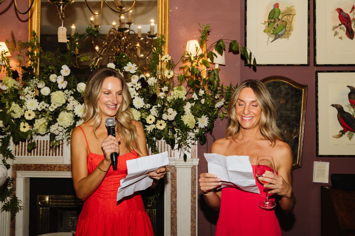 Two people in red dresses giving speeches at elegant event with floral decorations.