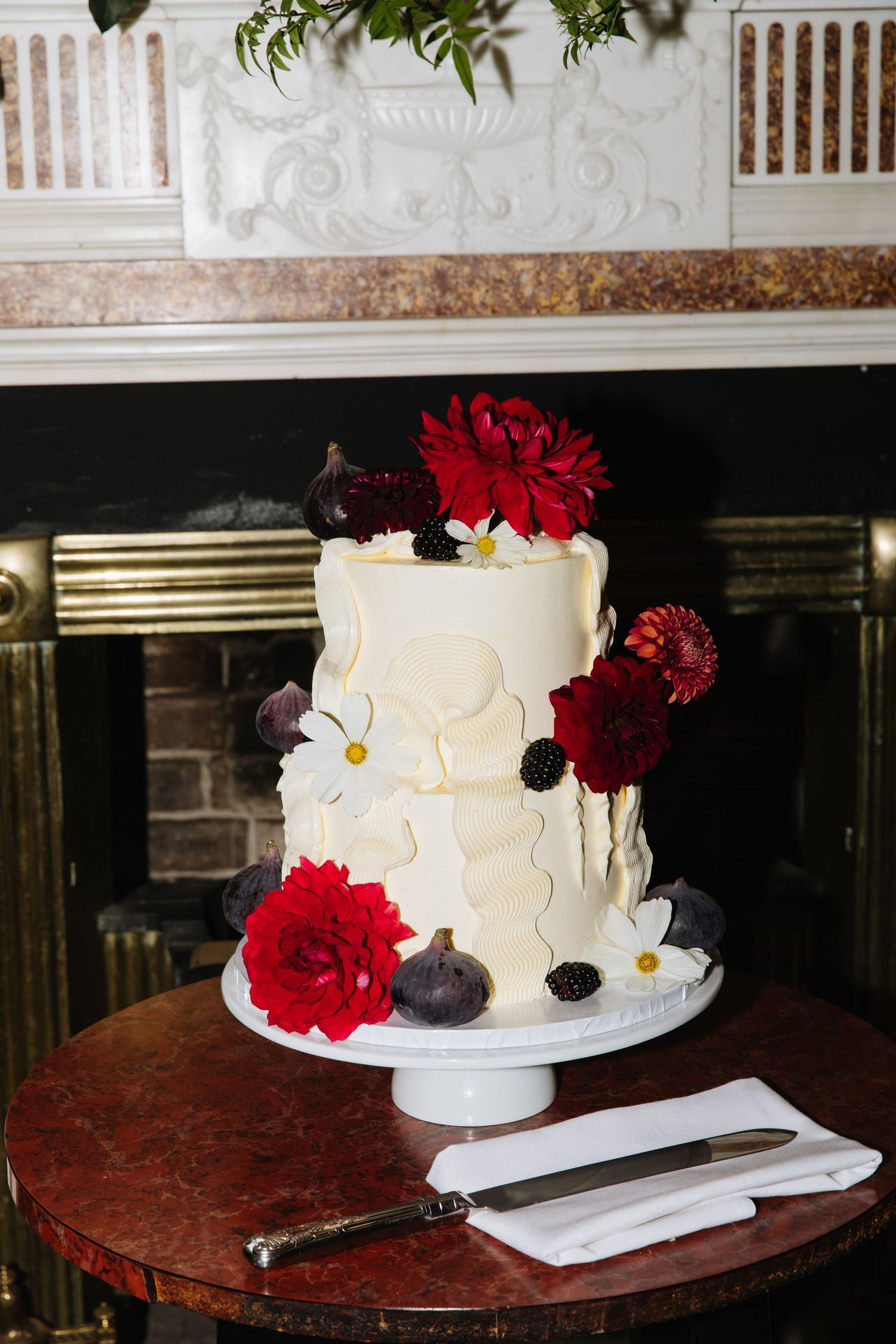 White tiered wedding cake decorated with red flowers and daisies on marble table.