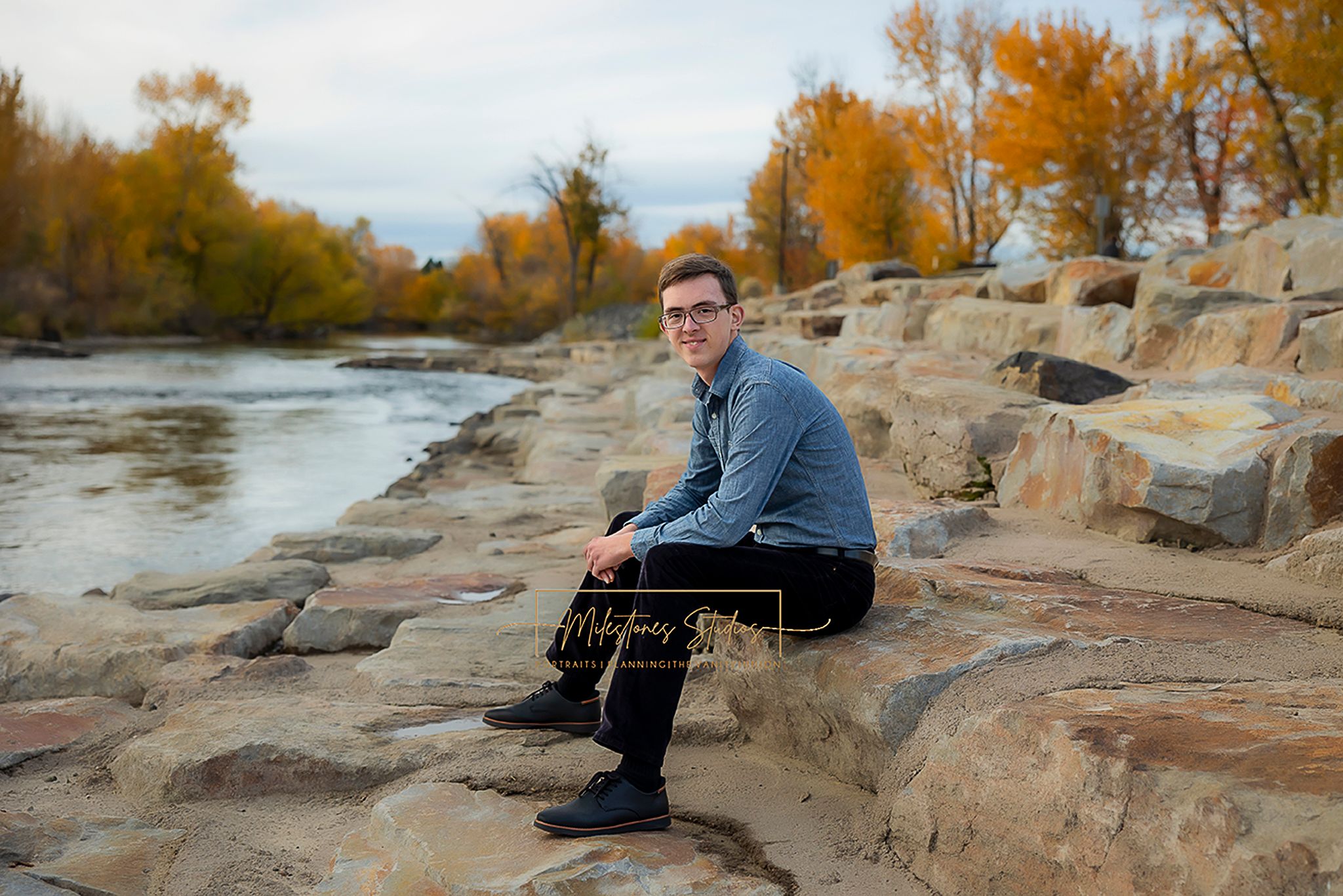 Boise Senior photos Boise white water park. Boy sitting on rocky riverbank in black jeans and blue shirt