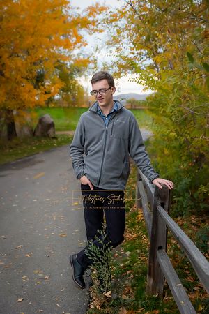 Boise Senior photos Boise white water park Boy standing near low fence in walk path surrounded by autumn trees