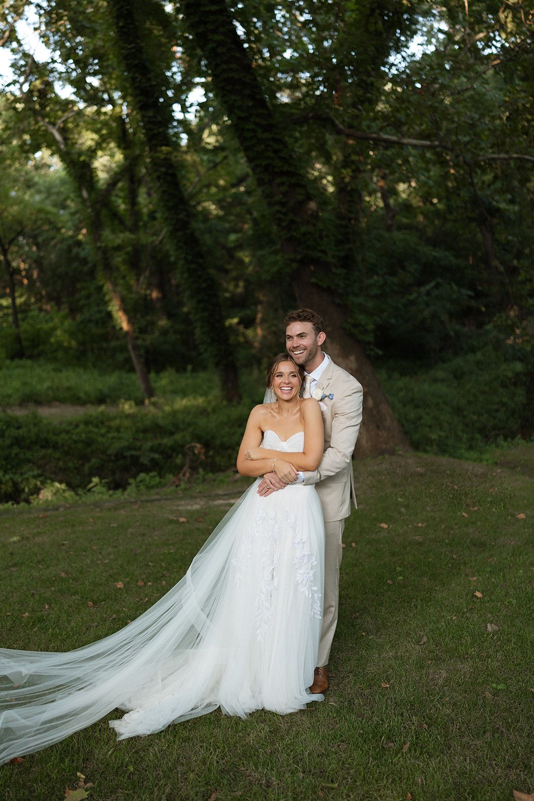 Wedding couple poses together on grassy field with flowing white dress and veil in natural outdoor setting.