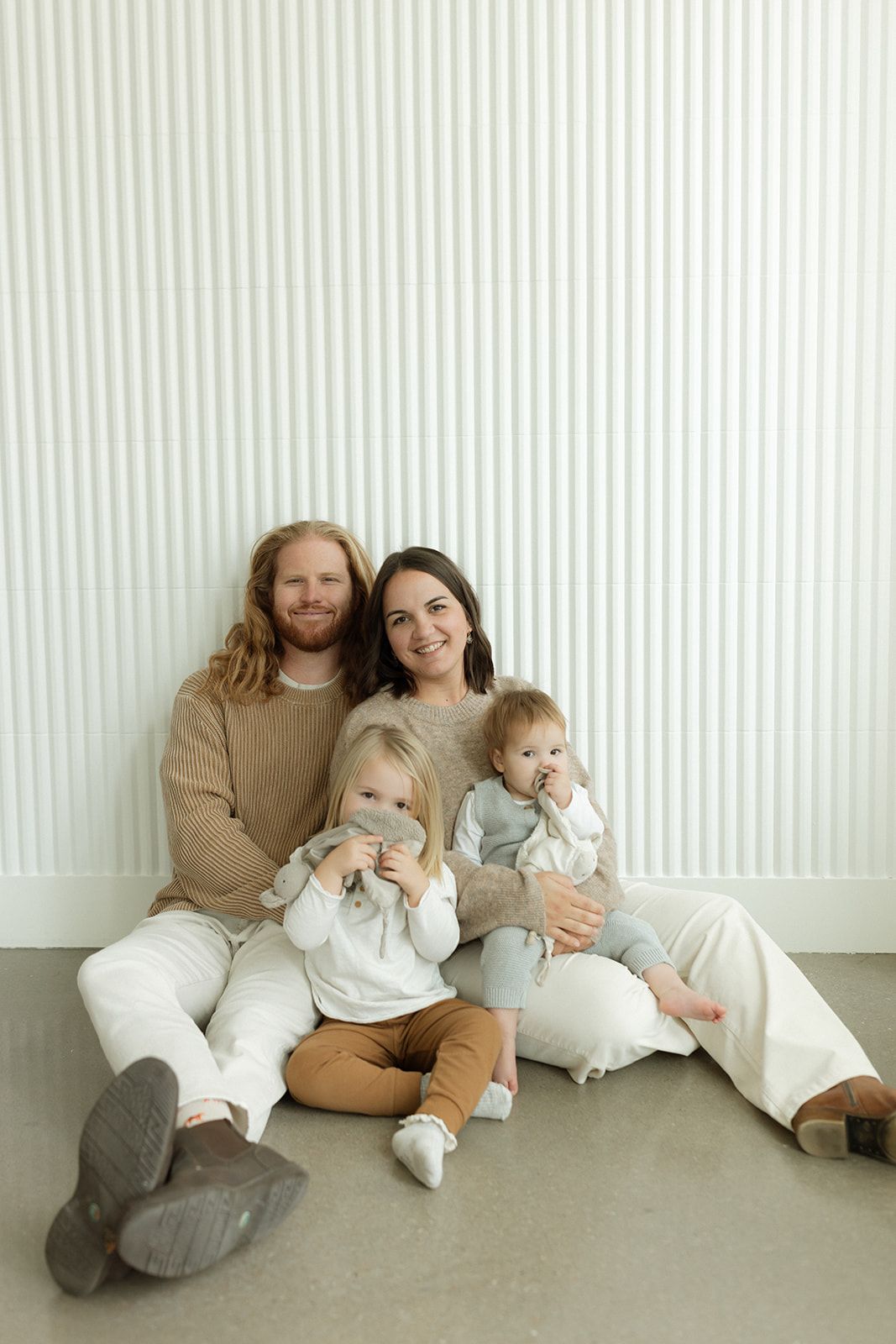 Family in coordinating cream and white outfits lounging together against white striped wall.