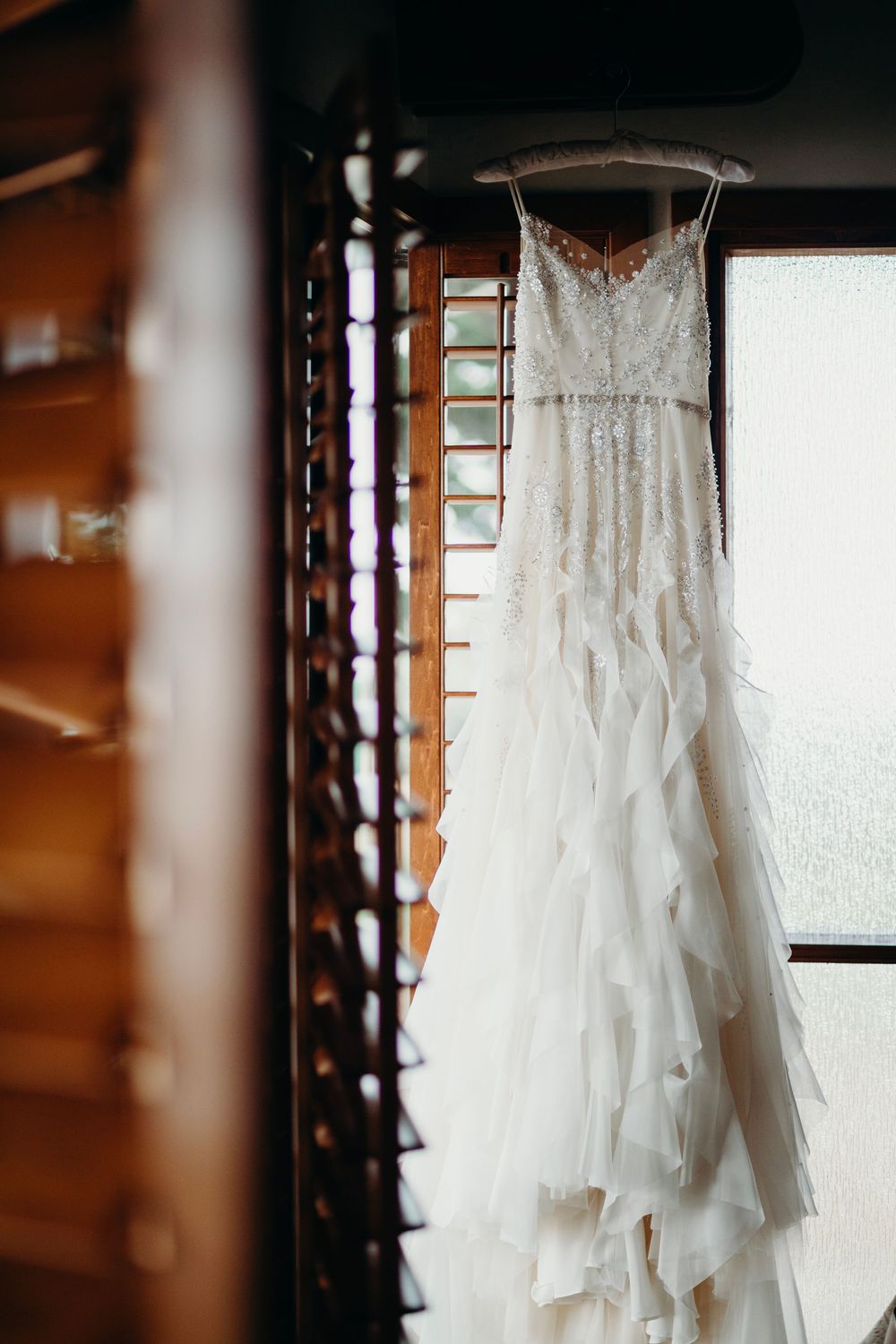 A white lace wedding dress with ruffled layers hangs elegantly against a wooden wall in natural lighting.