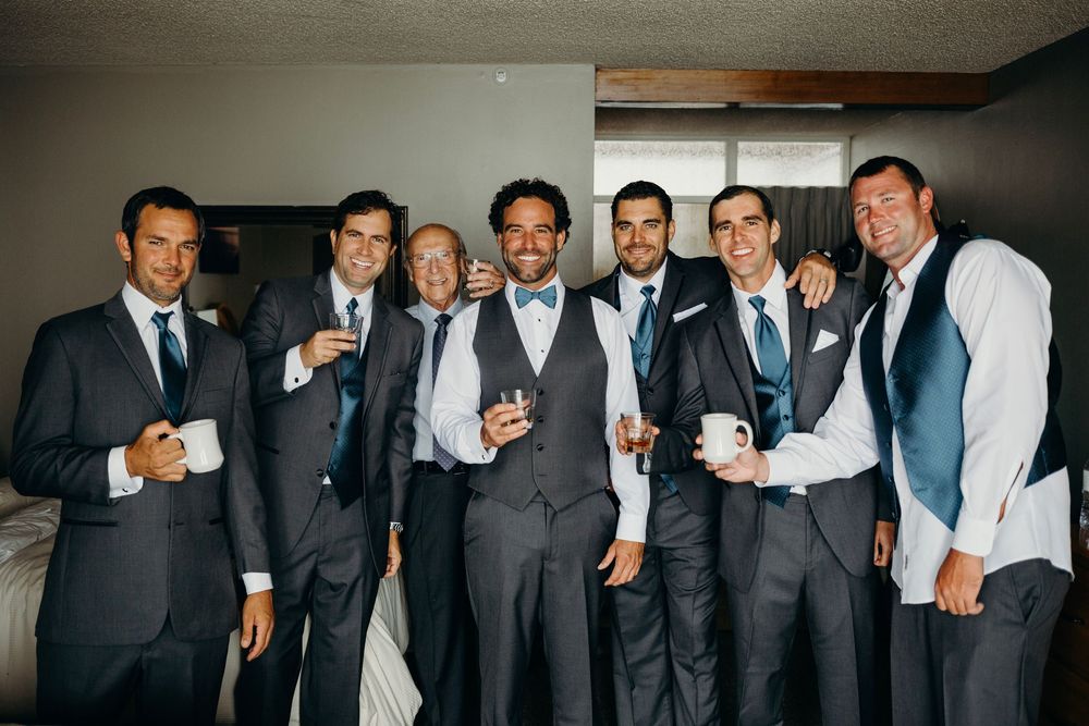 A group of groomsmen in suits and vests pose together for wedding party photos in a dimly lit room.