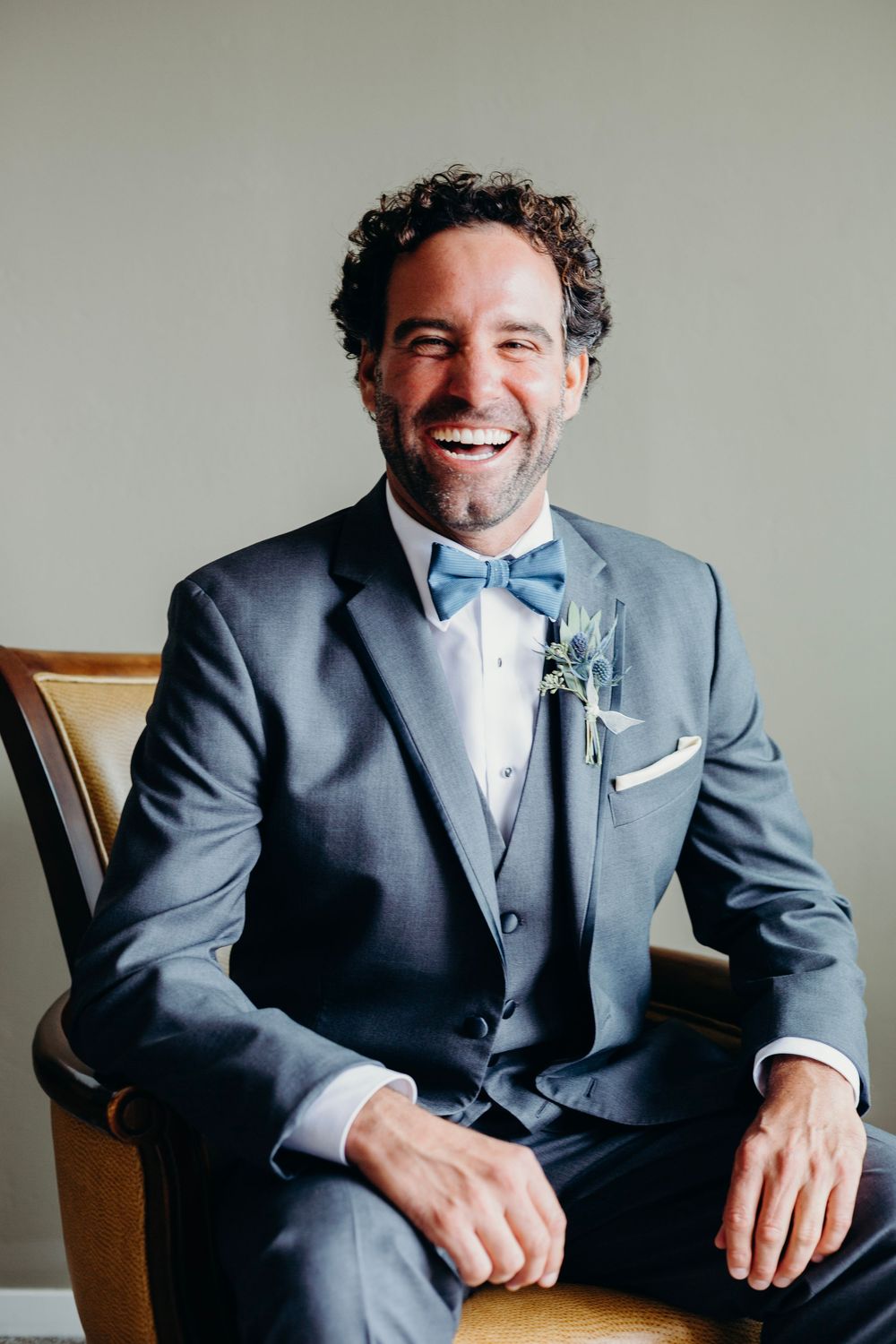 A groom in a charcoal grey suit and light blue bowtie smiles while sitting in a wooden chair.