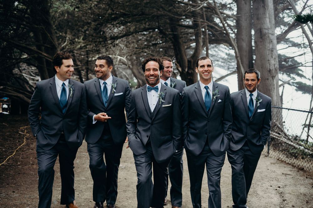 Groomsmen in matching navy suits share laughs while walking together on a misty outdoor path.