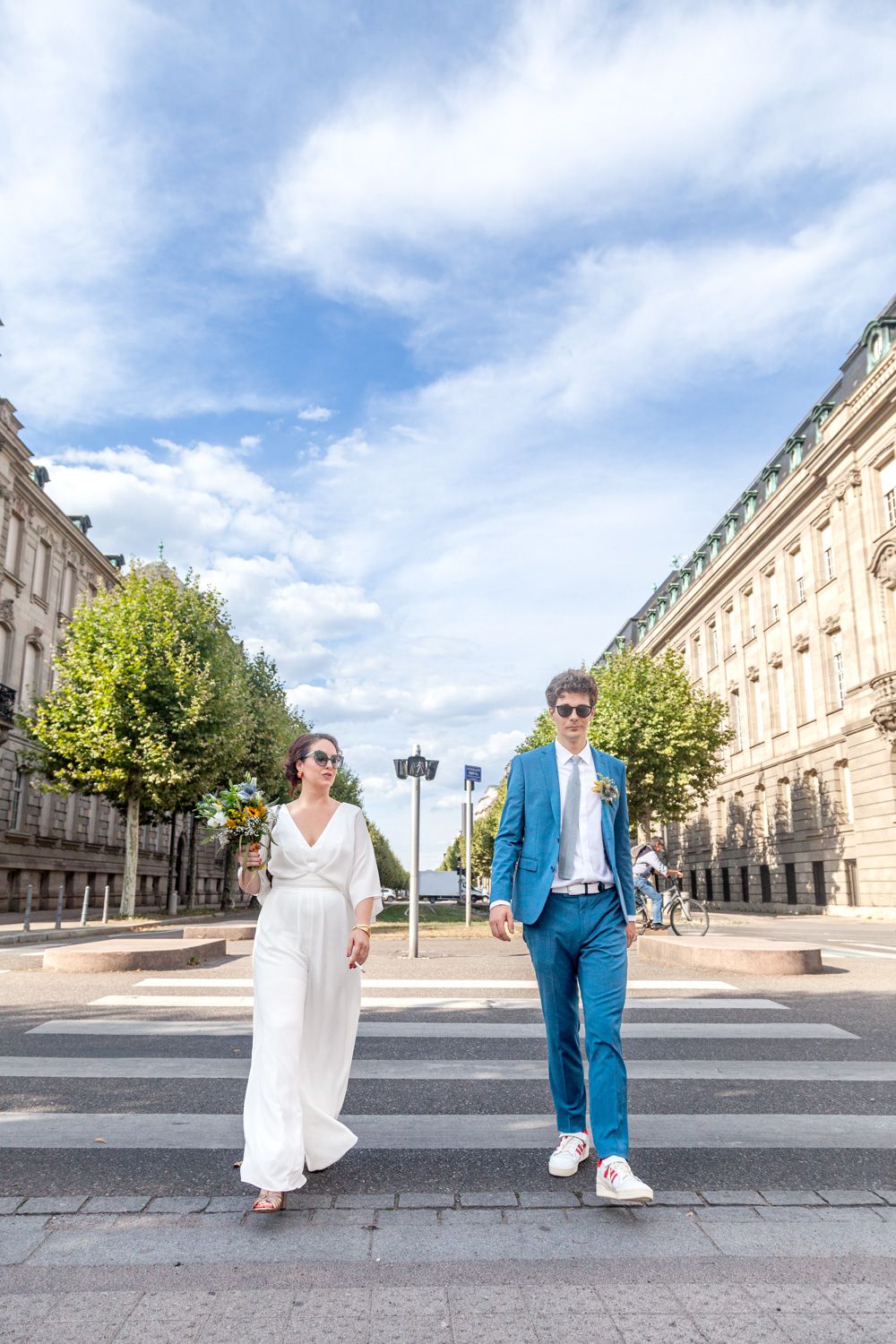 A couple poses in front of a historic building exterior, with the person in white sitting and blue suit standing.