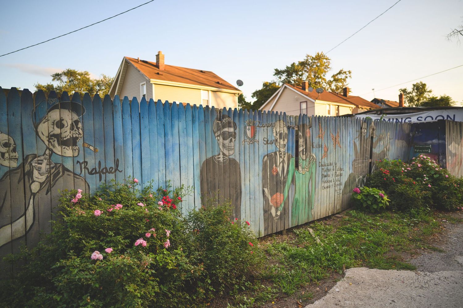 A rustic wooden fence stretches along a path with green foliage and houses visible in the background.