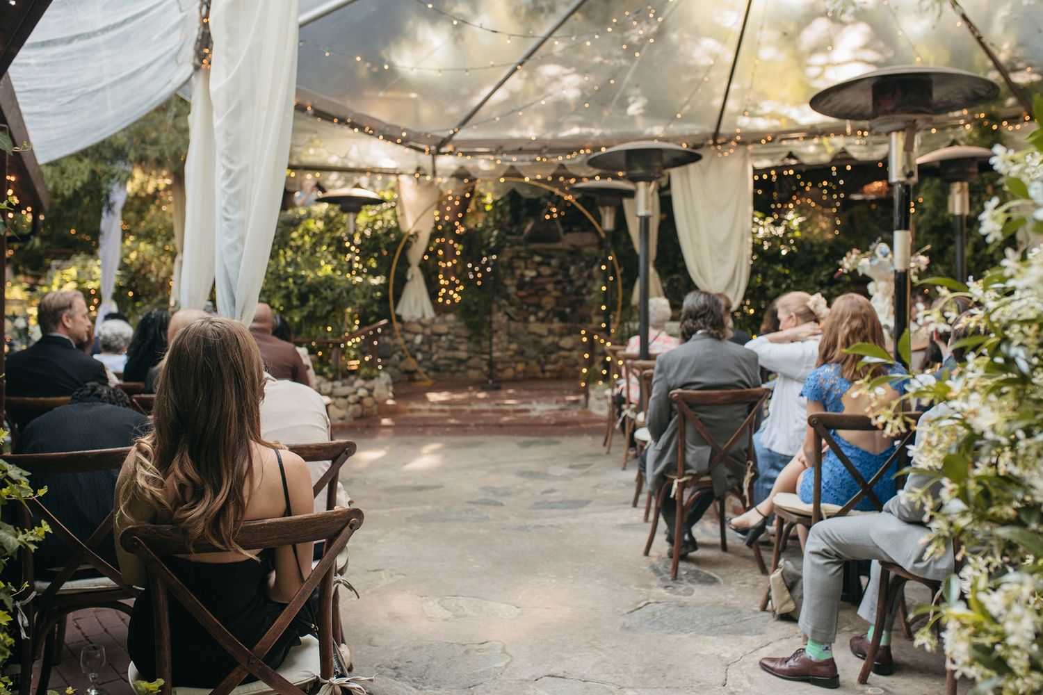 People enjoy a relaxing atmosphere at an outdoor wedding ceremony with string lights and greenery overhead.