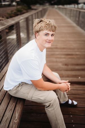 Person in white shirt and khaki pants sitting casually on wooden boardwalk in outdoor setting.