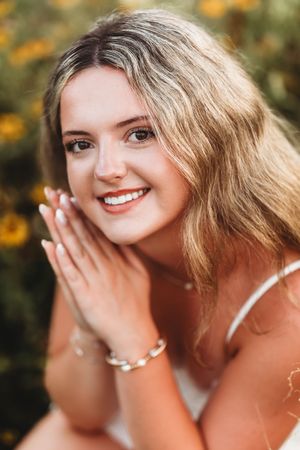 Smiling young woman with blonde hair and bracelet posing with hands together near yellow flowers outdoors.