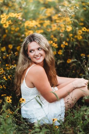 Smiling blonde girl in white dress sitting among yellow wildflowers in a sunny garden.