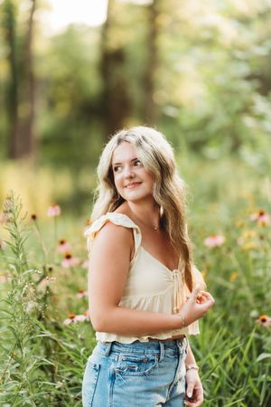 Young blonde woman in cream top and jeans smiling among wildflowers in a sunlit garden setting.