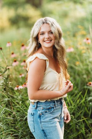 Smiling blonde teen girl in yellow top and jeans posing among wildflowers in a garden.