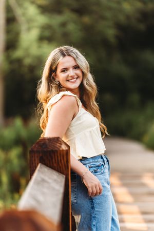 Happy blonde teenage girl in cream top and jeans leaning on wooden railing outdoors.