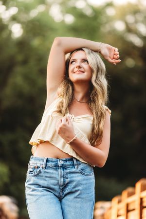 Young blonde woman in yellow crop top and jeans posing outdoors with arm raised, smiling in golden hour light.