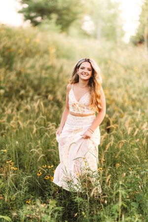 Smiling young woman in floral crop top and maxi skirt standing in a sunlit wildflower meadow.