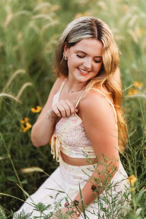Young woman smiling in a wildflower field, wearing a floral crop top and white skirt on a sunny day.