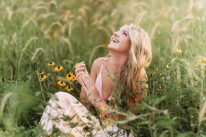 A joyful blonde woman sitting in a lush green field with yellow wildflowers, laughing and looking up on a sunny day.