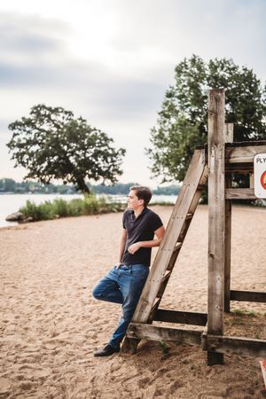 A person sits thoughtfully on a wooden beach structure while looking out over the sandy shore and water.