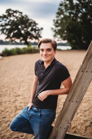 A casual outdoor portrait taken at a beach with wooden post, wearing dark polo shirt and jeans at sunset.