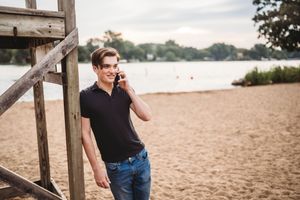 A person in a black polo shirt and jeans stands on a sandy beach near a wooden structure while talking on a phone.