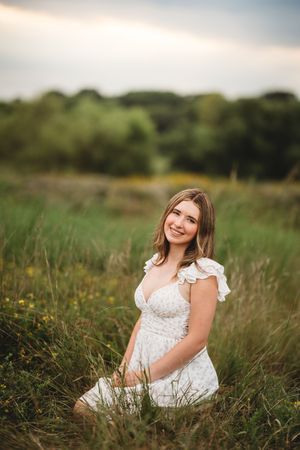 A person in a white lace dress sits gracefully in a sunlit meadow surrounded by tall grass and wildflowers.