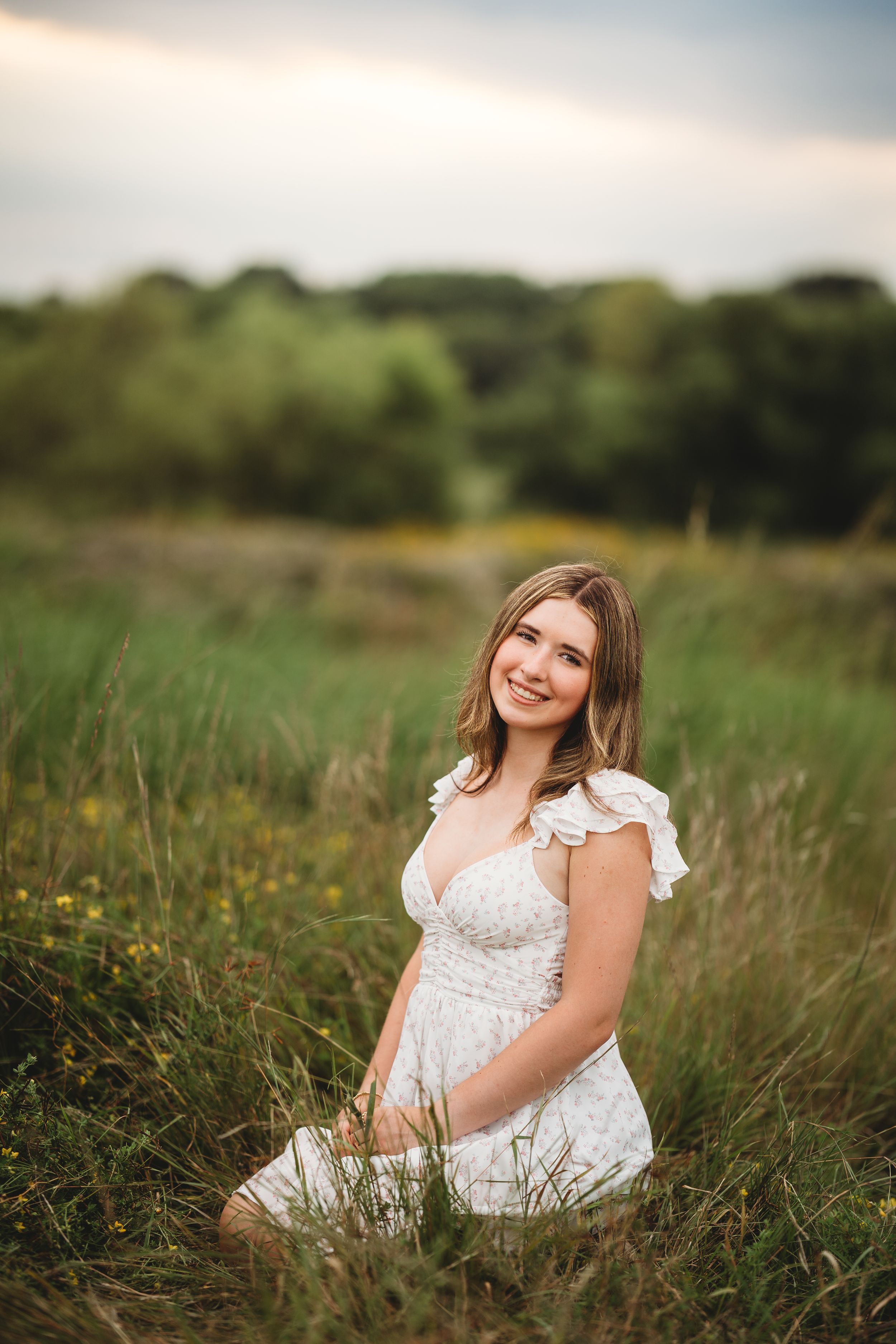 A person in a white lace dress sits gracefully in a sunlit meadow surrounded by tall grass and wildflowers.