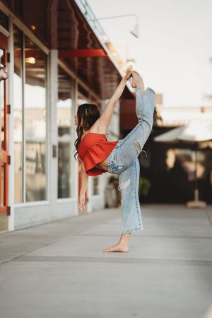Dancer in red shirt and jeans performs graceful high kick on sidewalk at sunset outside storefront.