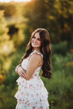 A person in a white floral dress poses outdoors during golden hour with sunlight creating a warm glow in the background.