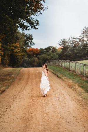 A figure in a white dress walks along a rustic dirt path surrounded by autumn trees and fencing in a rural setting.
